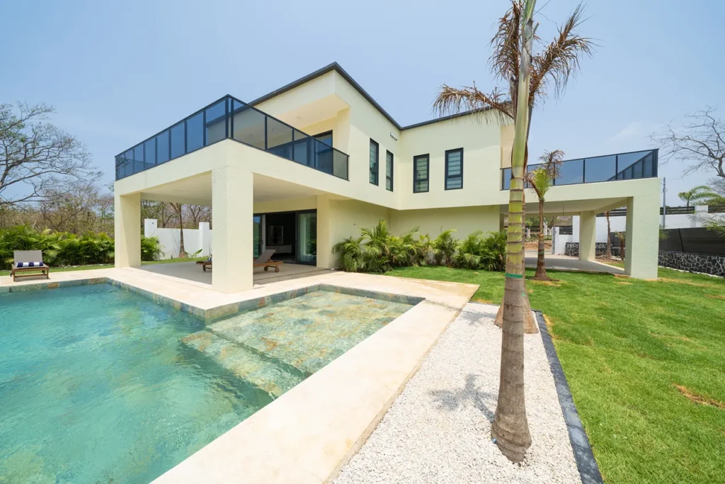 Casa de Acero's two-storey white facade and glass balcony beside a turquoise pool and palm tree.