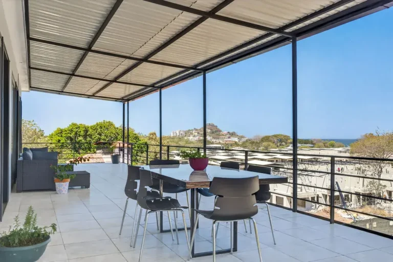 Balcony dining table under a steel roof with treetop and ocean views.
