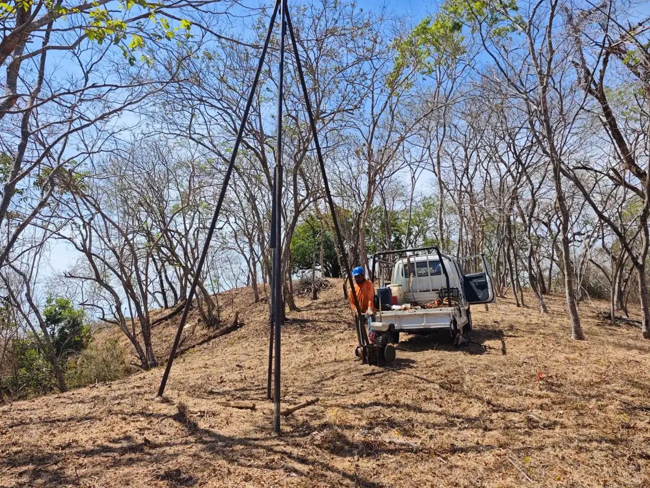Tripod drilling rig and a crew member in an orange shirt beside a white pickup truck on a hillside lot in Guanacaste