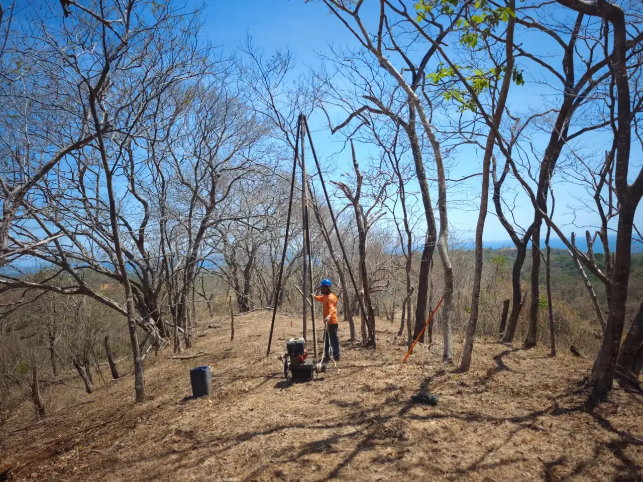 Crew member in an orange shirt and blue hard hat at a tripod drilling rig and engine on a dry hillside lot in Guanacaste, with the ocean in the distance
