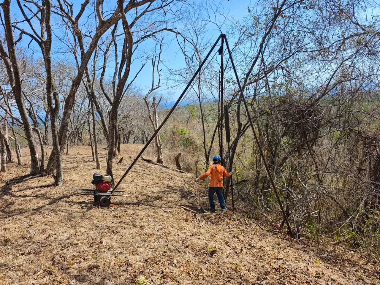 Tripod drilling rig on a dry forest hillside lot in Guanacaste, with a crew member in an orange shirt and blue hard hat and a portable engine