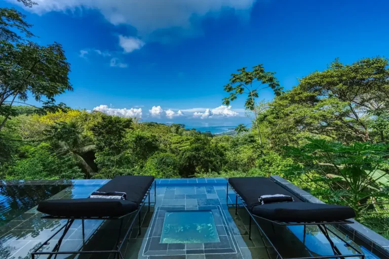 Infinity pool with black loungers set against blue sky and rainforest.